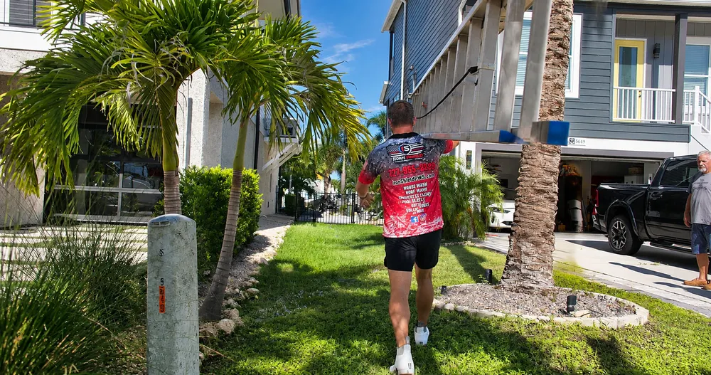 Iron Shield technician crossing a front lawn toward a two-storey Hudson home, carrying the cleaning line out to the awnings above