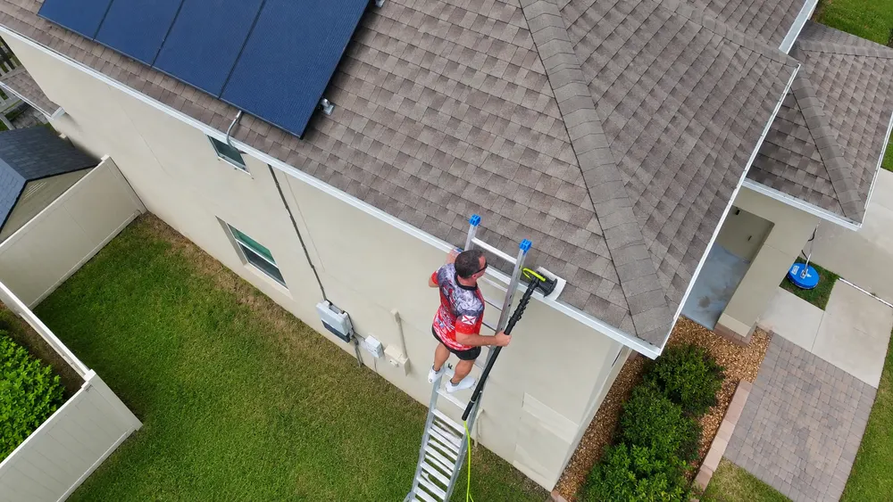 Iron Shield technician on an extension ladder, scrubbing the gutter and fascia line of a two-storey Hudson home