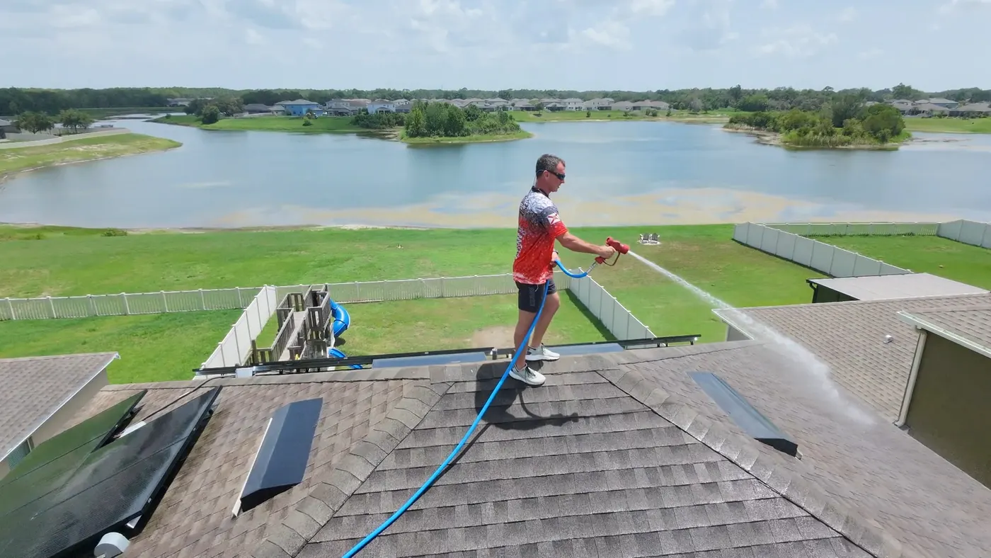 Iron Shield technician on the ridge of a shingle roof, soft washing around the solar panels with a lake behind the property