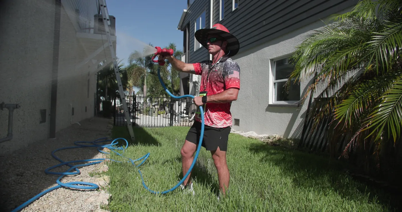 Iron Shield technician spraying an even arc of cleaning solution across the side wall of a Hudson home, hose running back across the lawn