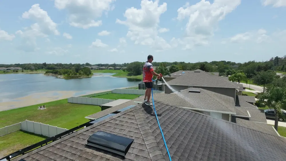 Iron Shield technician standing on a shingle roof, spraying a cleaning treatment across a home in an HOA community