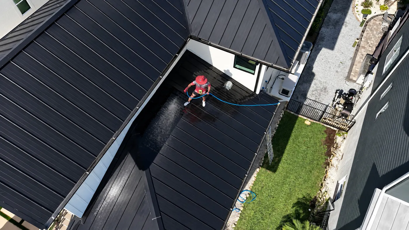 Top-down view of an Iron Shield technician washing a dark standing-seam metal roof with a hose-fed system