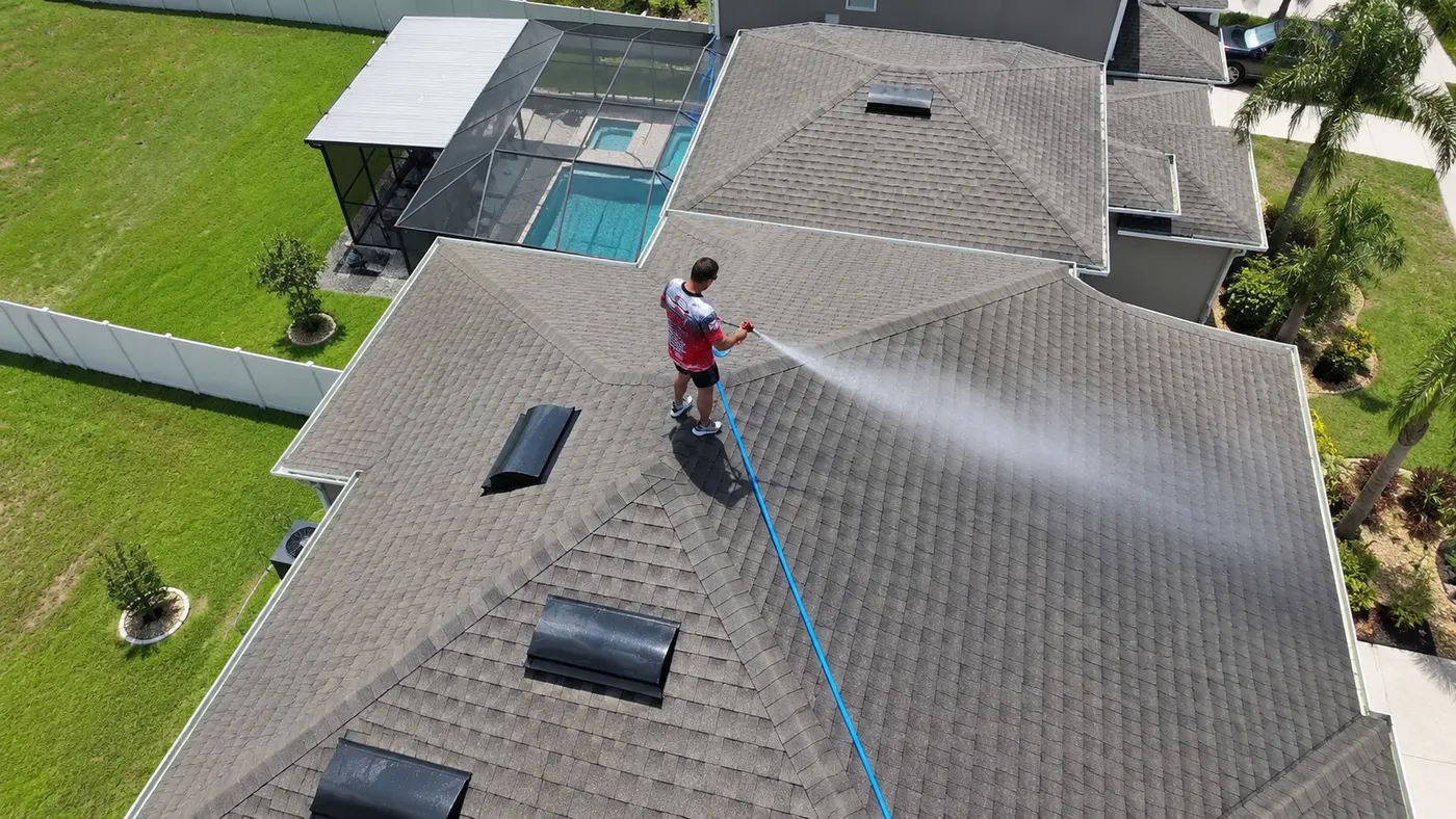 Aerial view of an Iron Shield technician applying a controlled cleaning treatment across a shingle roof beside a screened pool