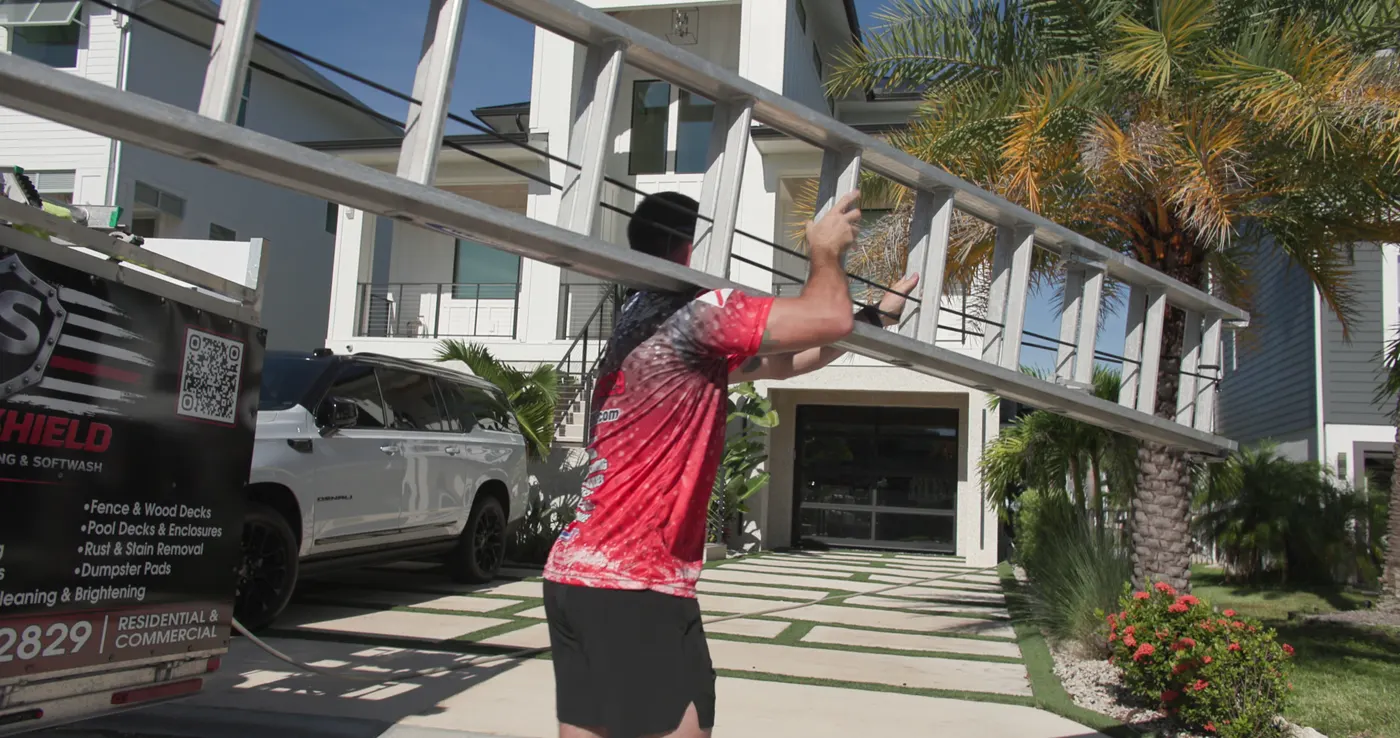Iron Shield technician at the front of a home, raising a long-reach pole toward the covered balcony above the entry
