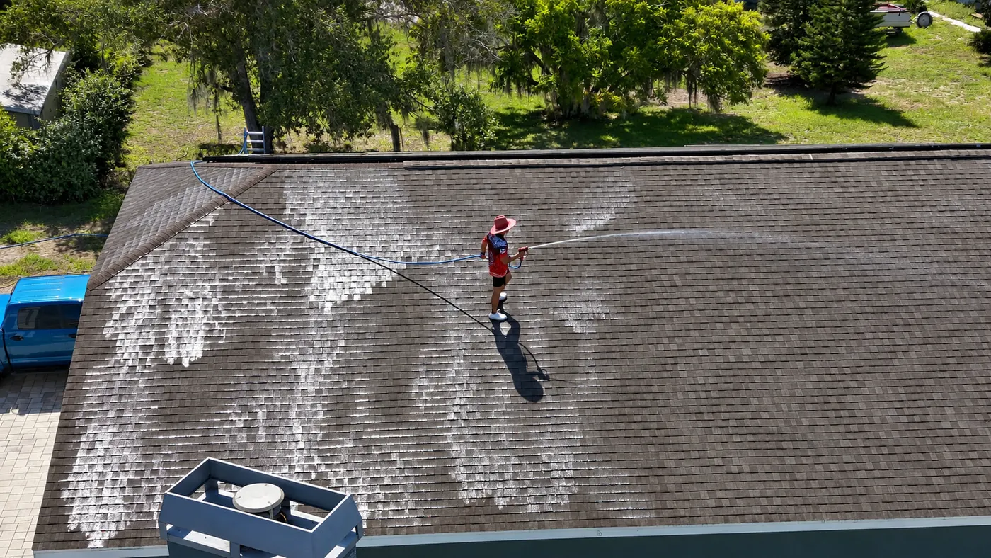 Aerial view of an Iron Shield technician rinsing a broad roof surface, the cleaned sections showing plainly against the untreated ones
