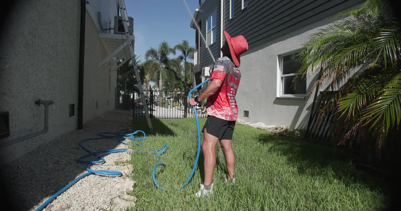 Iron Shield technician aiming a soft wash wand up at the upper wall and overhang of a two-storey home